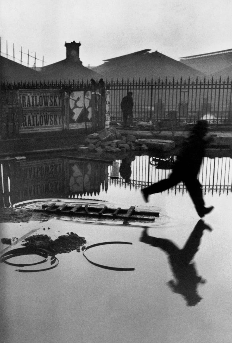 Auf dem Sprung, Paris, 1932: „Da war ein Zaun“, erzählte Henri Cartier-Bresson später, „und ich steckte meine Kamera durch den Zaun.“ Der springende Mann. Die Pfütze. Die Spiegelung. Und dieses Plakat im Hintergrund. 1932, hinter dem Bahnhof Saint-Lazare, Paris. „Es war der Bruchteil einer Sekunde“, sagte er. Das war seine Fotografie-Philosophie: der entscheidende Augenblick, in dem sich alles fügt.